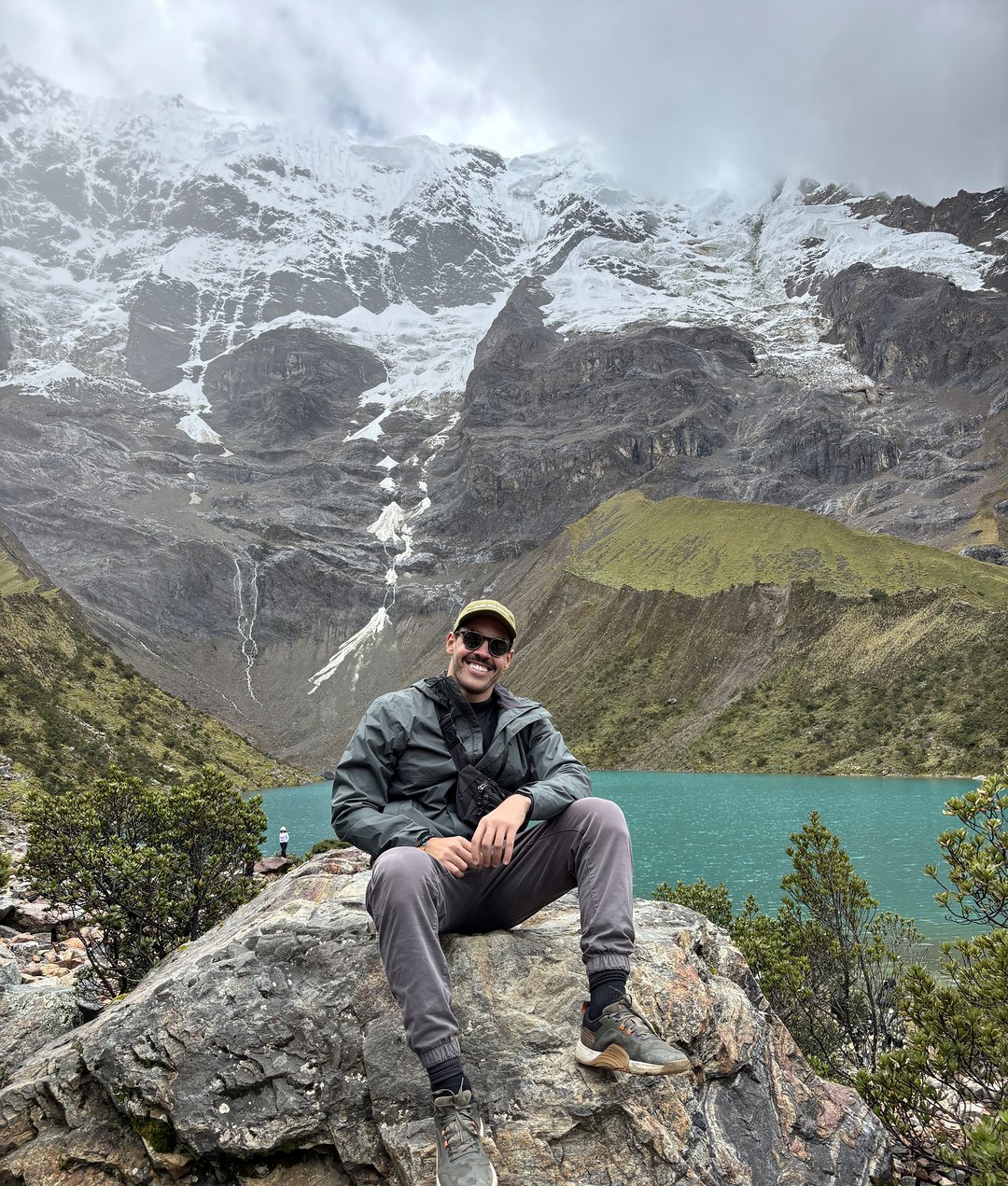 Lucas sentado numa pedra diante de uma lagoa turquesa, aos pés de montanhas nevadas nos Andes